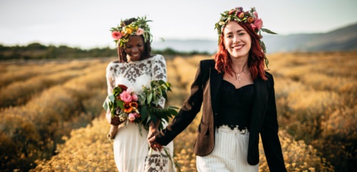 Black and white lesbian brides, holding hands, joyful walking in the yellow flower field in sunset
