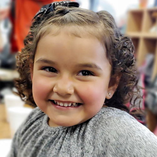 Young Kindergarten-age girl smiles while sitting at a table in classroom