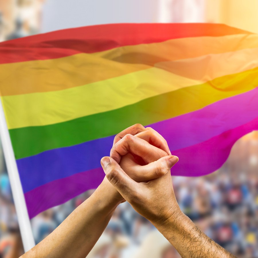 Cropped shot of a couple holding hands and wave in front of a rainbow flag flying on the sidelines of a summer gay pride parade